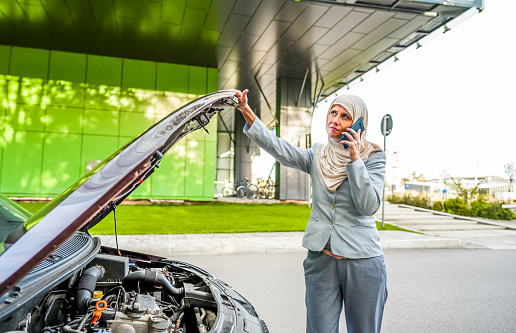 Mature Muslim businesswoman standing in front of a broken car and calling a car assistance service