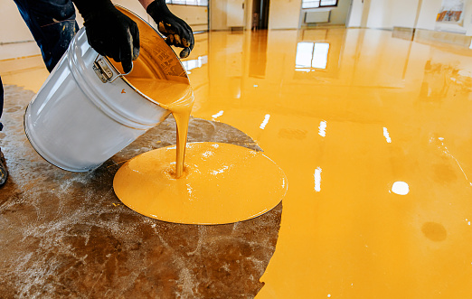 A male person in a yellow shirt and blue jeans pouring a clear liquid into a white bucket on an orange-colored floor