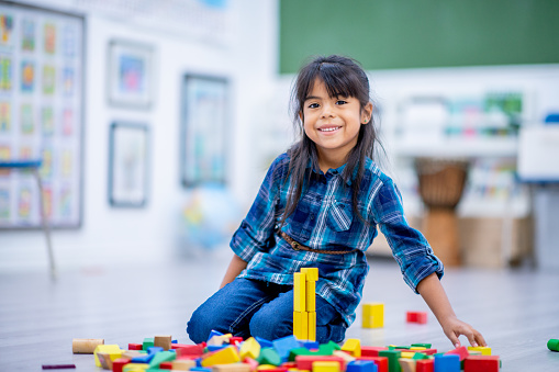 A Kindergarten student, of Hispanic decent, is seen playing on the floor with a pile of blocks during a free play time.  She is dressed casually and smiling.