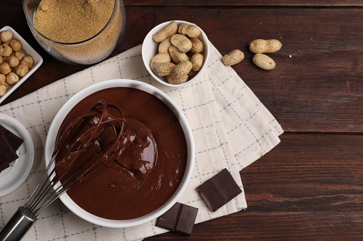 Bowl of chocolate cream with whisk and nuts on wooden table, flat lay. Space for text