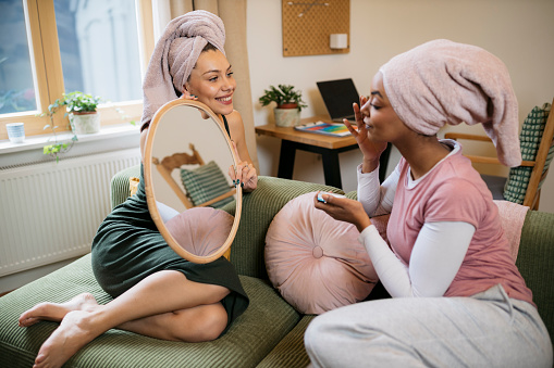 Pretty young woman holding a mirror for her friend applying face cream sitting on sofa at home after shower. Woman helping female friend in morning doing cosmetic procedure at home.