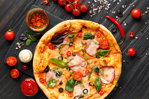 Pizza with jerky meat, ham, olives, cheese and herbs, sauces and fresh vegetables on a black stone plate on a dark wooden background, top view. Traditional Italian food.