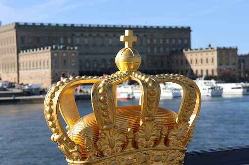 Stockholm Royal Palace. Photo from Skeppsholmsbron with a beautiful golden crown in the foreground. Blue sky. White archipelago boat on calm lake.