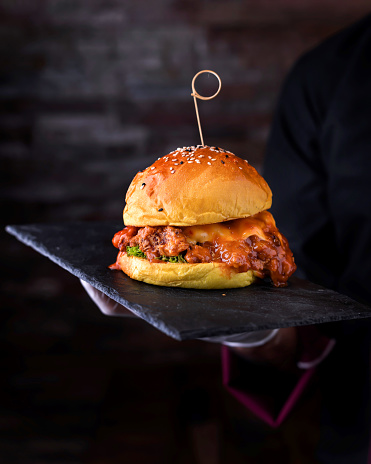Buffalo Chicken Burger with sauce and lettuce leaves served in cutting board holding in hand isolated on wooden table side view of arabic food