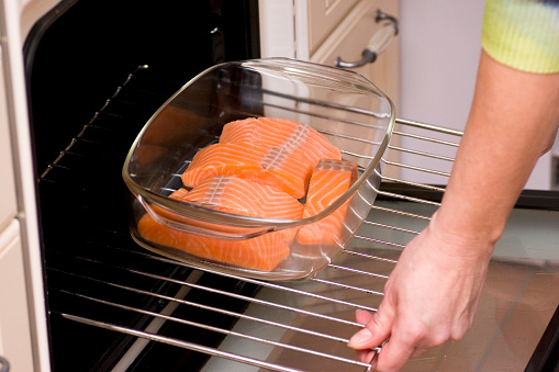 Women insert a glass casserole with fresh wild salmon fish filllets into the oven.