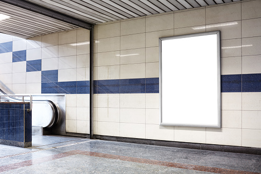 Blank billboard in a subway station wall. Wall is made of tiles and there is a blue coloured border in the middle. Billboard is oriented vertically and standing on the right side of frame. Billboard is empty so you can write or add something on it. - Clipping path of billboard included.