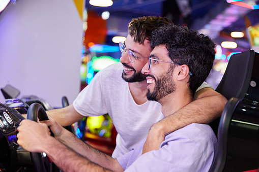 Happy gay couple playing arcade driving simulator. They are together hugging with a loving attitude.