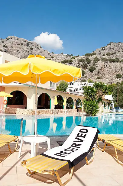 A beach towel with the word 'Reserved' spread onto an empty sun lounger beside a swimming pool