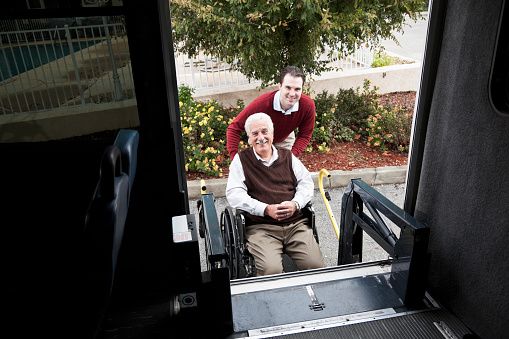 Young man (20s) helping senior man (60s) onto shuttle bus equipped with wheelchair lift.