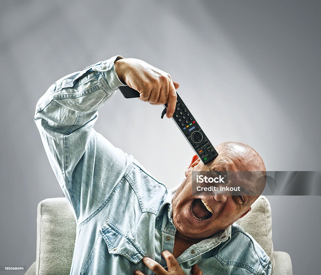 TV Madness isolated old man pointing a tv remote control to his head. HeA's screaming and wears a blue jean shirt. Adult Stock Photo