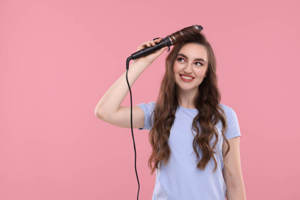 istock Beautiful young woman using curling hair iron on pink background, space for text 1850705707