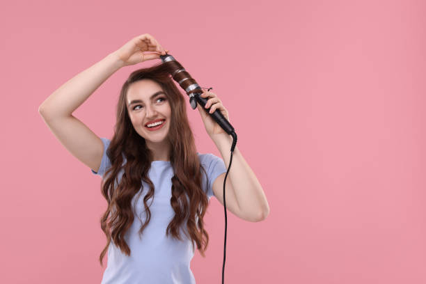 istock Beautiful young woman using curling hair iron on pink background, space for text 1850706235