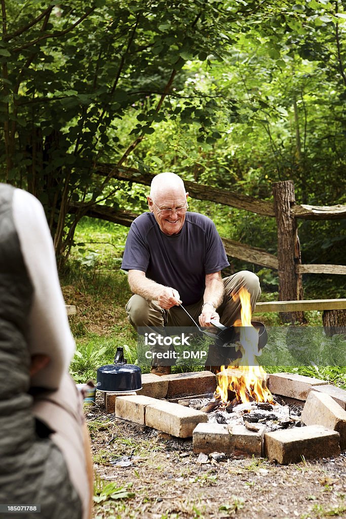 Homem velho feliz cozinhar na fogueira de acampamento - Foto de stock de Fogueira - Fogo ao Ar Livre royalty-free