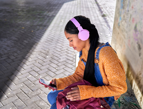 Gorgeous digital nomad Colombian Latina girl sitting on a bench waiting for the bus and listening music and checks bus schedules at the city bus station in her smart phone