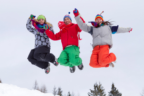 Three smiling young people in multi colored winter clothes jumping downhill in snow.
