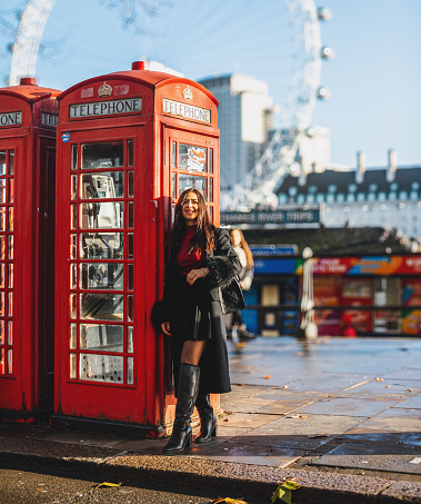 Portrait of a  happy, stylish, and smiling young woman posing while looking at camera and standing by the iconic red telephone booths in London. Her poised stance against this famous backdrop captures the essence of contemporary elegance. Ideal for conveying a blend of style and iconic landmarks, this stock photo is perfect for fashion blogs, lifestyle magazines, and social media content looking to showcase the charm of London's iconic streets. Iconic London, Stylish, Smiling, Young woman, Portrait, Red telephone booths, Famous backdrop, Contemporary elegance, Style, Landmarks, Fashion blogs, Lifestyle, Social media