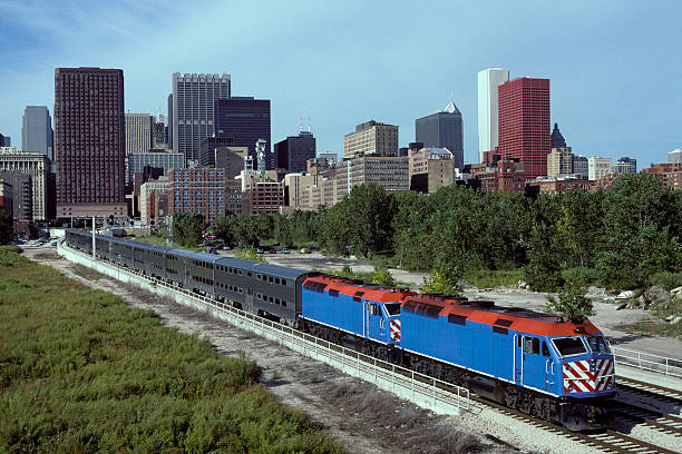 Two streamlined blue diesel locomotives depart busy city with a train of stainless steel double-decker commuter carriages. Train passing left to right in front of cityscape of skyscrapers. The locomotives have hi-visibility safety stripes on the front.  Logos and numbers removed.