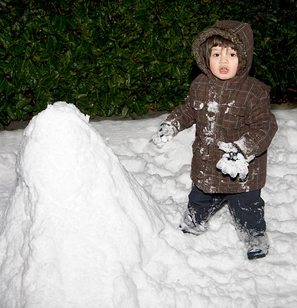 Toddler having fun building a snowman in the garden and getting his hands and feet covered in snow.