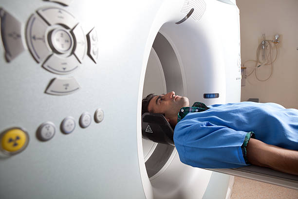 Adult Man Having Medical Examination Via CT Scanner Young man in blue protective hospital clothes having medical examination via CAT scan.The focus is on model.The photo is taken with a full frame DSLR camera in horizontal composition.The man is in his thirties and has brown hair located on the right of frame. pet scan photos stock pictures, royalty-free photos & images
