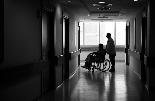 Silhouette of man on his wheelchair in dark corridor.Male Nurse is pushing the chair.The image was taken in real hospital.Light at the end of corridor is visible for symbolic purpose.Black and white tone is used for effective atmosphere.Photo was shot with DSLR camera.