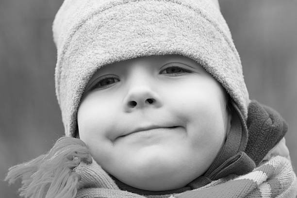 Young child wearing a beanie and winter coat, smiling broadly and enjoying a playful indoor activity in black and white. Scanned from analog film