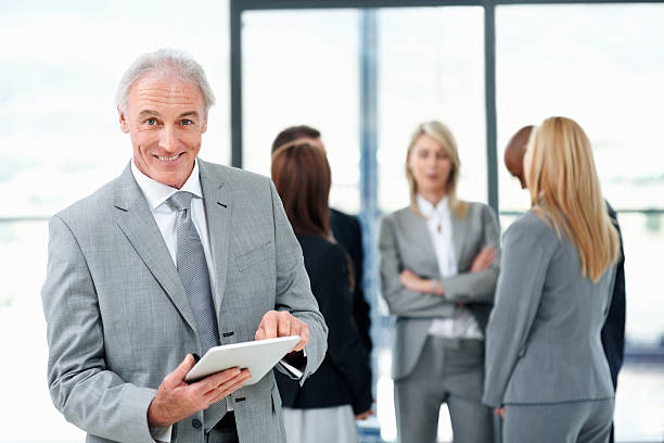 Happy business man using tablet pc Mature business man smiling and using tablet pc with colleagues in background peoples alliance for democracy stock pictures, royalty-free photos & images