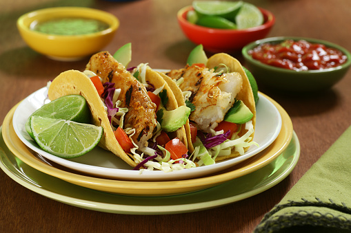High-resolution digital capture of a plate of fresh fish tacos made with crispy grilled fish, avocado, cabbage, tomatoes, and fresh lime, on golden corn tortillas. Dish is photographed on a warm wooden surface. Small bowls of salsa verde, lime slices, and  red salsa are visible in the background.