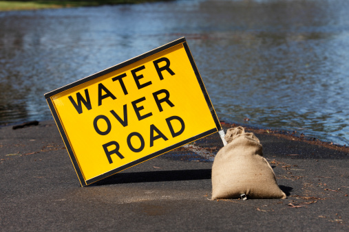 A yellow hazard sign with black text, warns of water over the road ahead.  Behind the sign water rises from a flooding river and advances towards to sign.  The road is flooded and impassable.