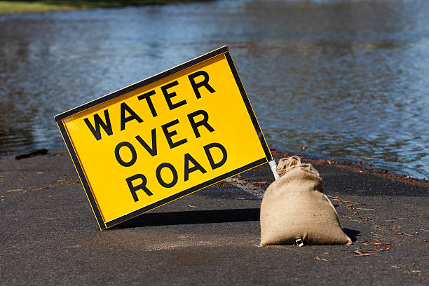 Yellow “Water Over Road” hazard sign and flooded street A yellow hazard sign with black text, warns of water over the road ahead.  Behind the sign water rises from a flooding river and advances towards to sign.  The road is flooded and impassable. road closed sign horizontal road nobody stock pictures, royalty-free photos & images