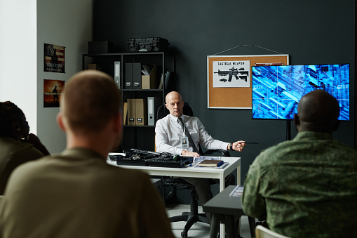 Confident mature officer sitting by workplace and making presentation of military tactics on interactive screen to group of students