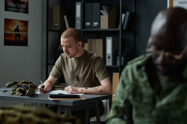 Young man in t-shirt sitting by desk and making notes in copybook