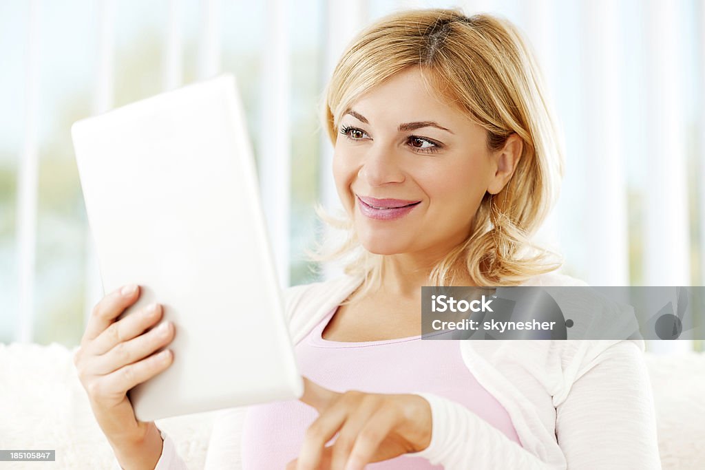 Beautiful woman using a digital tablet. Cheerful blonde woman relaxing at home and using her touchpad. Adult Stock Photo