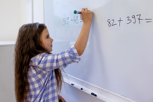 Little schoolgirl counting math equation on whiteboard in school.