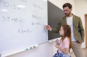 School teacher helping pupil writing on white board solving math problem in classroom.
