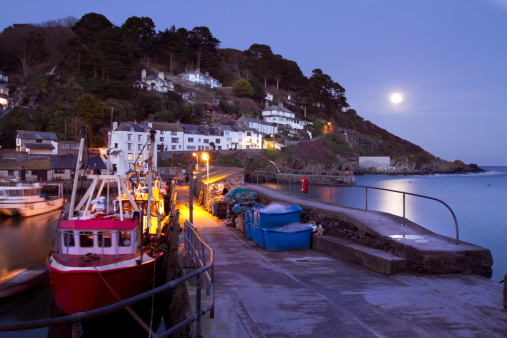 A tranquil scene of Polperro harbour at night. The picture show the moon rising over the sea and the pier of the harbour with fishing boats moored against it.The image was captured with a long exposure creating some motion blur in the boats and ocean.