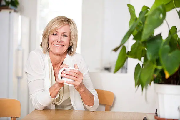 Happy, middle aged woman drinking hot tea in her white kitchen. She is smiling at the camera. Woman is in her 50s and has both hands around the coffee mug. She is very genuine and warm. Green plan in of the right of the image.  
