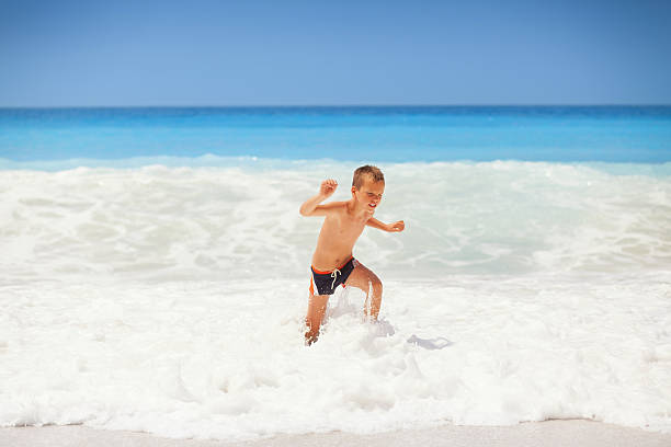 Young little boy on the beach Little boy enjoying the beautiful beach, smiling and running in front of large wave. Photo was taken on one of the most beautiful beaches in the world. The Egremni beach. Lefkada island in Greece. egremni beach lefkada island greece stock pictures, royalty-free photos & images