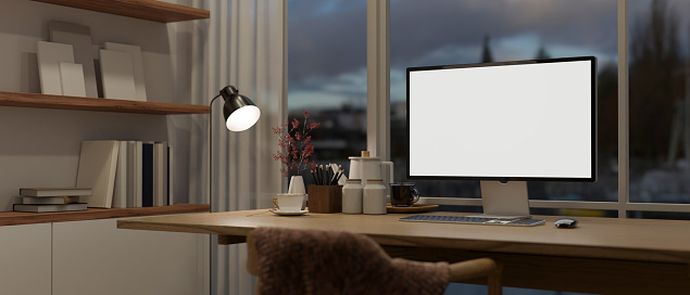 Interior design of a modern, cosy home office workspace at night with a PC computer mockup on a wooden desk against the window, a floor lamp, a wooden chair, and a shelf. 3d render, 3d illustration