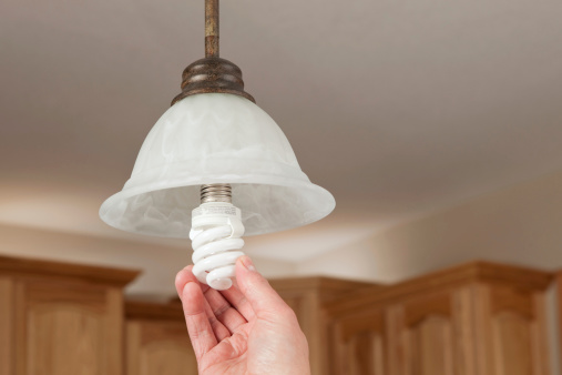 A male hand is installing an energy efficient compact fluorescent light bulb (CFL) into a ceiling hung pendant fixture. The background is kitchen cabinets with above cabinet lighting.