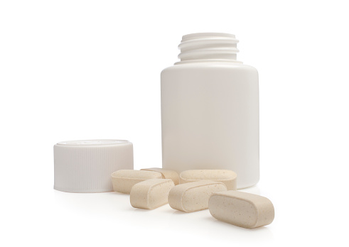 A studio shot of some pills scattered in front of an open pill container on a white background with reflections