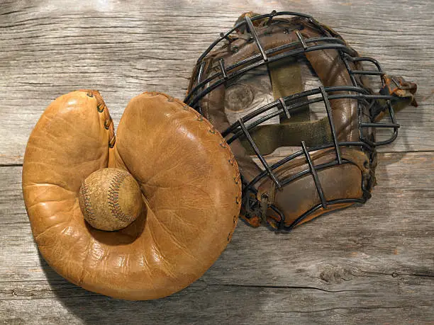 An antique catcher's mitt and mask, with an old baseball on a weathered wood background.