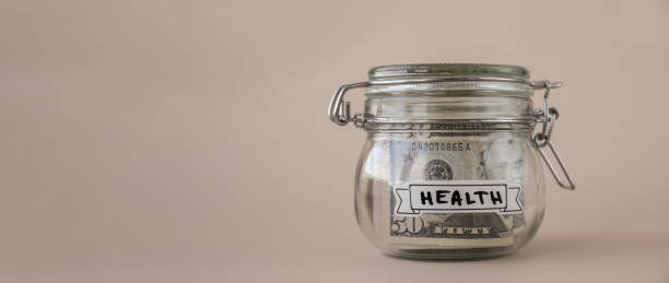 Saving Money In Glass Jar filled with Dollars banknotes. HEALTH transcription in front of jar. Managing personal finances extra income for future insecurity Saving Money In Glass Jar filled with Dollars banknotes. HEALTH transcription in front of jar. Managing personal finances extra income for future insecurity medical transcription stock pictures, royalty-free photos & images