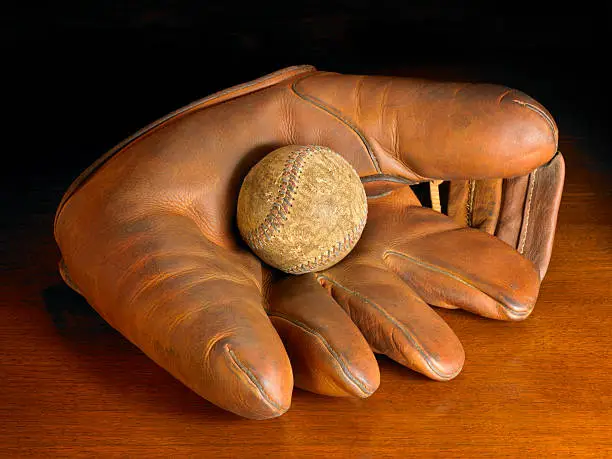 An antique baseball glove and baseball on a wood background. Warmly lit for a nostalgic feeling.
