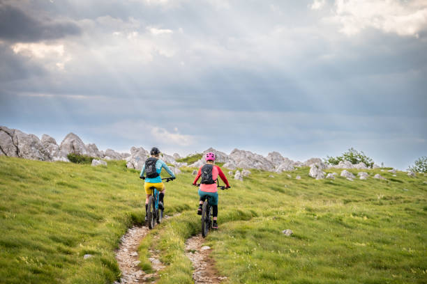 Two women riding electric mountain bikes together at green meadows landscape full length back view