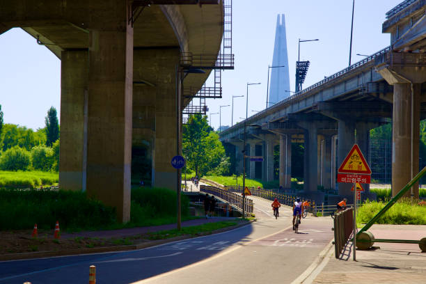 Seoul, South Korea - June 2, 2023: Cyclists traverse a bridge over Tan Stream with the soaring Olympic Boulevard overhead, and the iconic Lotte World Tower rising majestically in the background.