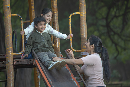 Cheerful young mother assisting son and daughter in playing on slide at outdoor playground during weekend
