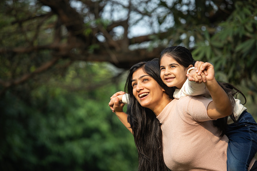 Cheerful young mother with arms outstretched piggybacking cute smiling daughter in park during weekend
