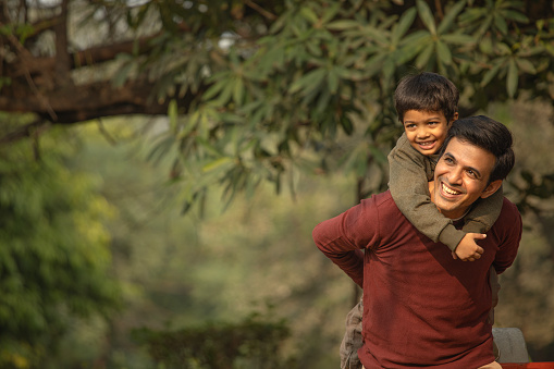 Smiling young father piggybacking cute playful son and looking away while standing against trees in park during weekend