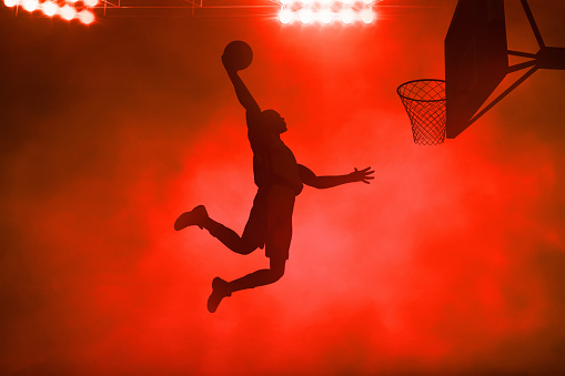 3d illustration shadow silhouette of young professional basketball player slam dunk on dark red smoke background