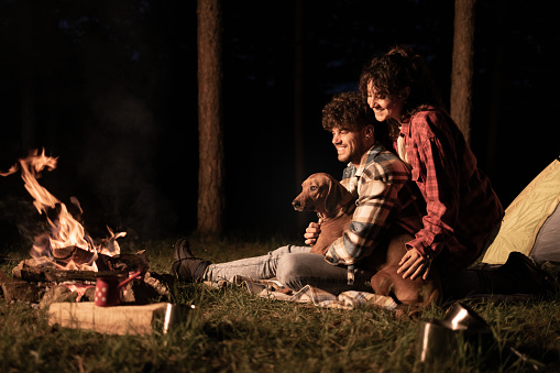 Happy couple and their dog relaxing by the fire during camping night in nature. Copy space.